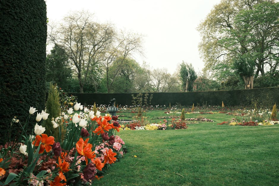 A well-maintained garden featuring a lush green lawn bordered by colorful flower beds with tulips, hyacinths, and other seasonal blooms in shades of white, orange, pink, and purple. On the left, a den